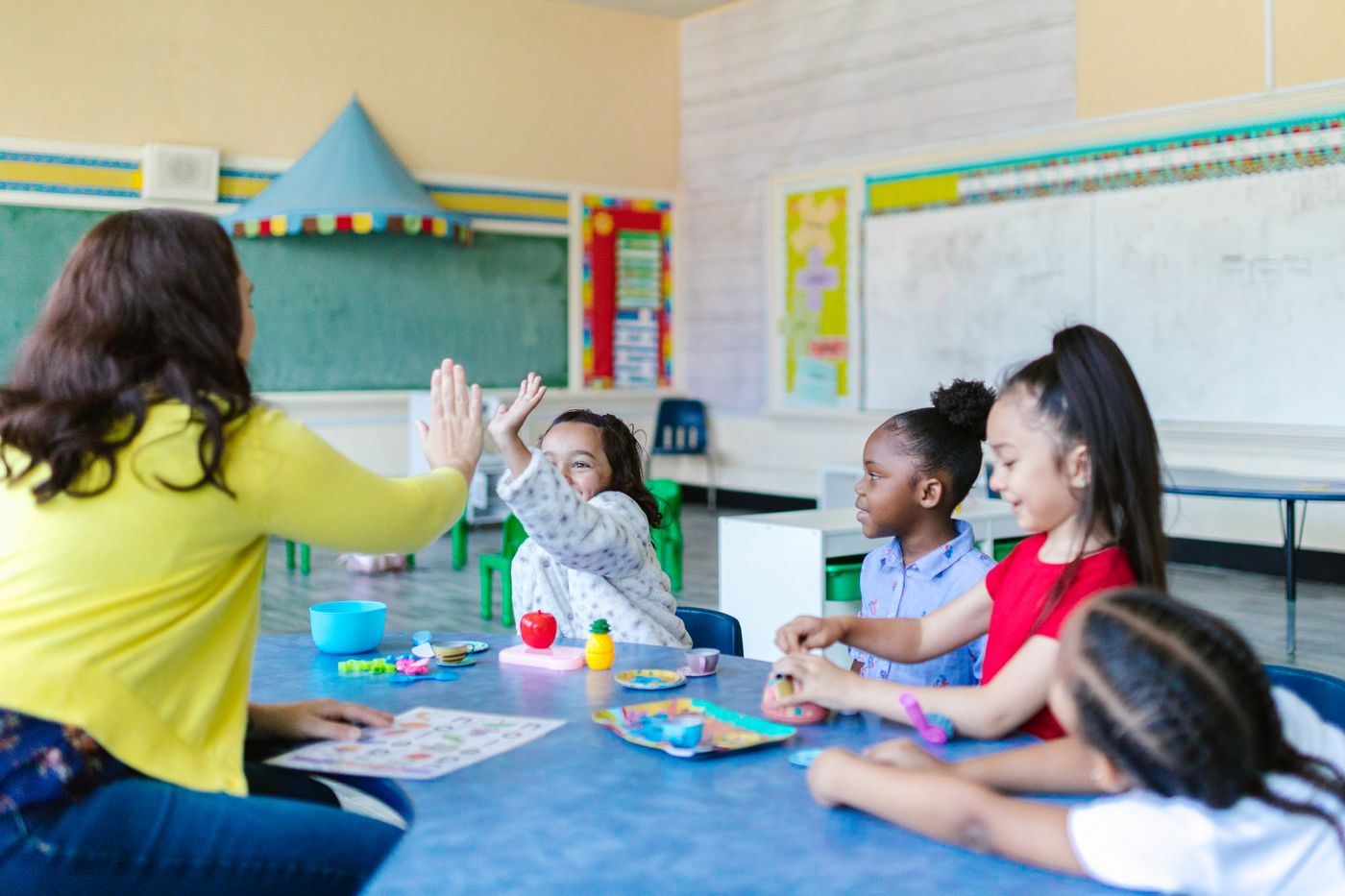 A teacher high-fives a student as children work on a hands-on activity at a shared table