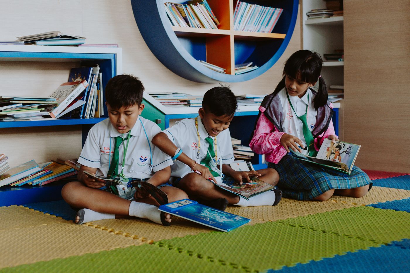 Three children sit together on a classroom rug, reading picture books